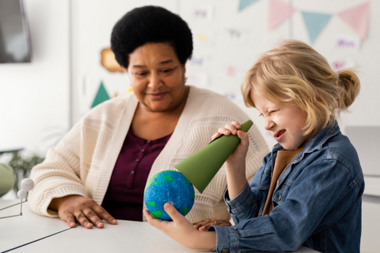 The image shows a teacher and child engaging in a classroom activity. The child is holding a green cone near a globe model, looking through it, while the teacher observes and smiles.