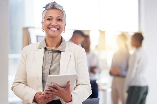 A person standing in a bright office environment, smiling and holding a tablet. In the blurred background, several colleagues are having a discussion in small groups.