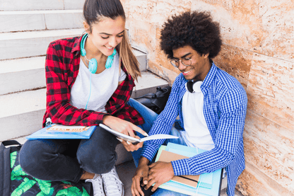 Two students sit on steps outside, smiling and looking at an open book together. One holds folders and wears headphones around their neck; the other points at the book and has teal headphones around their neck. Both appear relaxed and engaged in conversation or study.