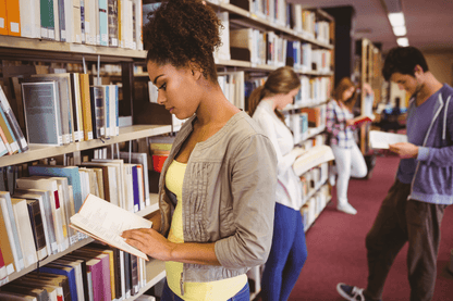 A group of students stands in a library aisle, each focused on reading a book. Shelves filled with books line both sides, and the setting appears quiet and studious. The individuals are spaced out, absorbed in their reading.