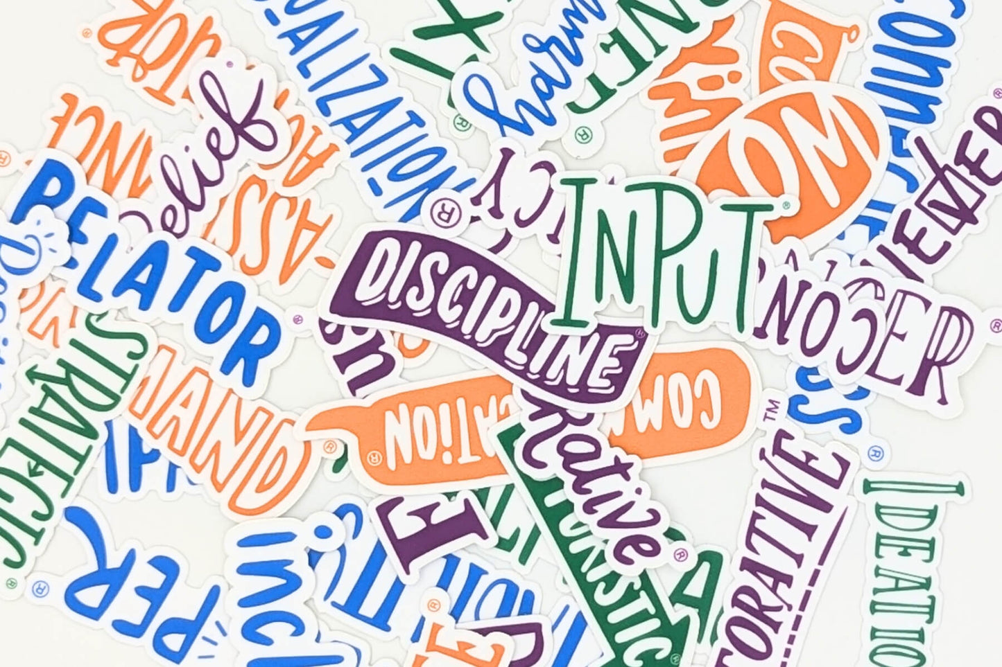 Close-up of colorful word stickers with terms such as 'Input,' 'Discipline,' 'Relator,' 'Strategy,' and 'Belief,' scattered on a white surface. Stickers are based on the Gallup CliftonStregths® and colored based on the Clifton Strength Domain.