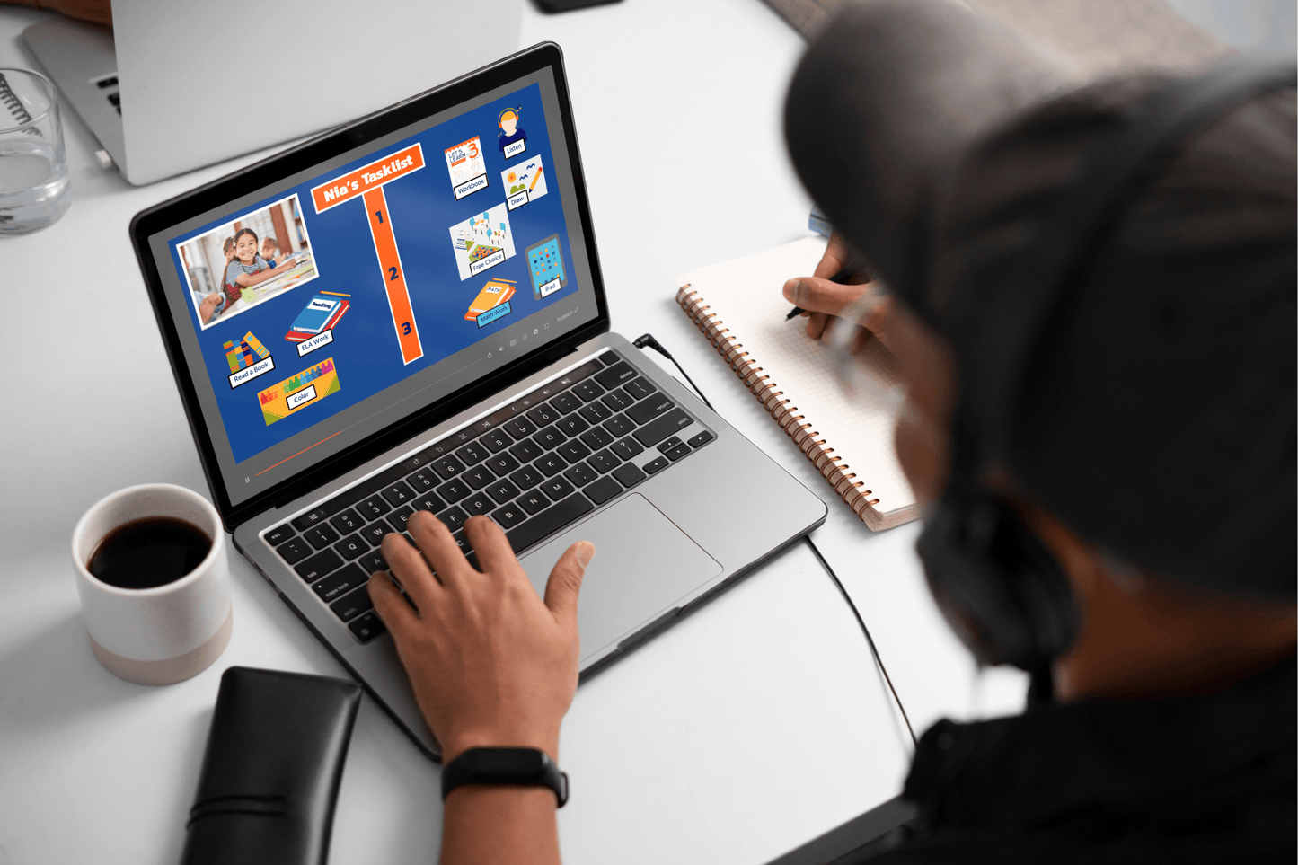 A person wearing headphones works on a laptop displaying a colorful project checklist. They write in a notebook, with a cup of coffee and a wallet nearby on a white table.