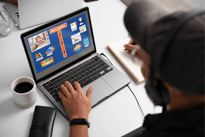 A person wearing headphones works on a laptop displaying a colorful project checklist. They write in a notebook, with a cup of coffee and a wallet nearby on a white table.