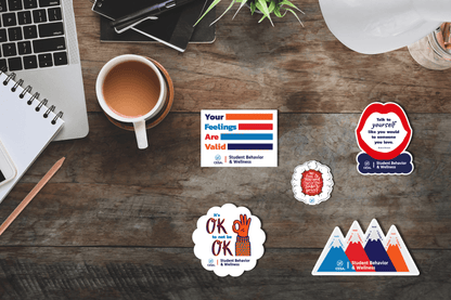 ticker set promoting student behavior and wellness displayed on a wooden desktop beside a laptop, notebook, coffee cup, and writing tools. Stickers include motivational phrases such as 'Your feelings are valid,' 'It’s OK to not be OK,' and 'Talk to yourself like you would to someone you love.' 