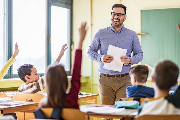 The image shows a teacher standing in front of a classroom, smiling and holding papers. Several students have their hands raised, engaged in the lesson.