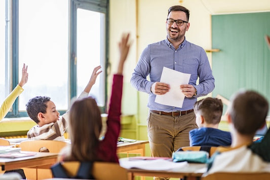 The image shows a teacher standing in front of a classroom, smiling and holding papers. Several students have their hands raised, engaged in the lesson.