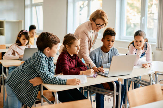 A teacher leans over to assist a small group of elementary students gathered around a laptop at their desks. The children are engaged, with some writing in notebooks while others watch the screen, and additional students work independently in the background.