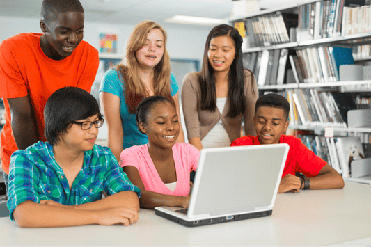 A group of students gathers around a laptop at a library table, smiling and engaged while looking at the screen. Bookshelves filled with books are visible in the background.