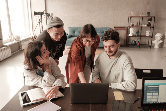 Four adults sit together at a table, collaborating around a laptop. One person points at the screen while the others watch attentively. Open notebooks, a smartphone, and another laptop are on the table, emphasizing teamwork and problem-solving in a modern workspace.