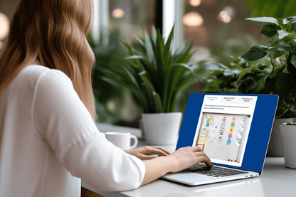 A woman with wavy hair works on a laptop at a desk with green plants. The screen displays colorful graphics. 