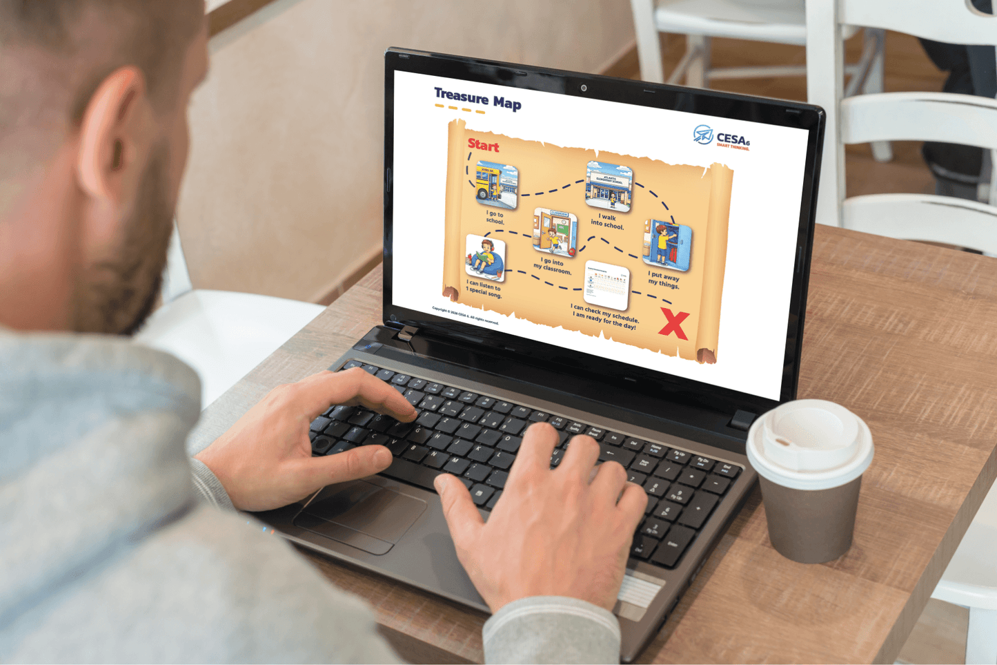 A person is using a laptop displaying a treasure map graphic. The background is a café setting. A paper cup is beside the laptop on the wooden table.