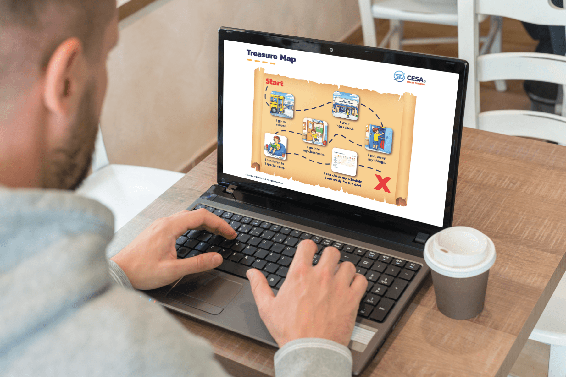 A person is using a laptop displaying a treasure map graphic. The background is a café setting. A paper cup is beside the laptop on the wooden table.