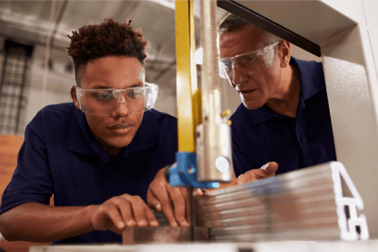 A student wearing safety glasses carefully operates a metal-cutting machine under the close supervision of an instructor, who is also wearing safety glasses. Both are focused on the task, emphasizing hands-on learning and safety in a career and technical education setting.