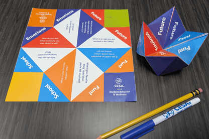 A colorful paper fortune teller, designed by CESA6 Student Behavior & Wellness, encourages children to engage in reflective conversations through fun and meaningful questions about emotions, school, the future, and favorite activities. The unfolded version shows vibrant color-coded sections with questions, while the folded version stands partially open. A pencil and a CESA6-branded marker lie nearby on a dark wooden surface.