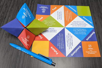 A colorful paper fortune teller from CESA6 Student Behavior & Wellness encourages meaningful conversations with thought-provoking questions about fun, relationships, emotions, and the future. A folded version sits next to an unfolded sheet with vibrant color-coded sections. A blue CESA-branded pen rests nearby on a dark wooden surface.