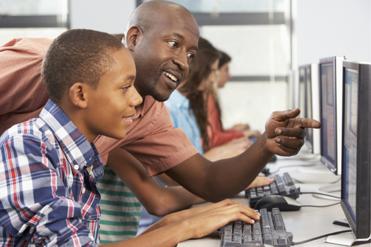 An educator leans in to guide a student at a computer, pointing at the monitor with a smile as the student types. Several other students work at computers in the background.