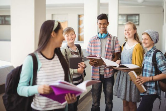 A group of students stands together in a school hallway, holding books and notebooks while smiling and talking. One student in the foreground walks toward them carrying folders, appearing engaged in the interaction.